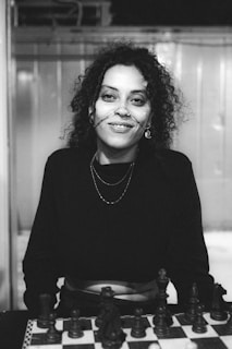 A person with curly hair and wearing a nose chain is smiling. They are dressed in a dark top and are seated behind a chessboard. The background appears to be an indoor setting with a metallic or reflective surface.