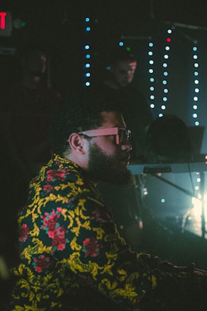 A man wearing glasses and a colorful patterned jacket with floral designs sits in a dimly lit environment. Behind him, a person works with electronic equipment, possibly DJ or sound engineering setup. The background is adorned with vertical strings of blue lights, creating a vibrant and artistic atmosphere.