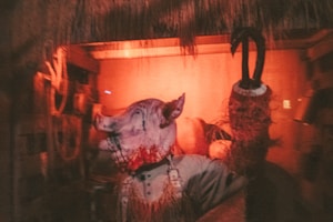 A dimly lit scene with an eerie atmosphere featuring a figure wearing a grotesque pig mask and a bloodied appearance. The setting appears to be rustic, perhaps resembling an old barn or shack, with a reddish-orange hue dominating the lighting.