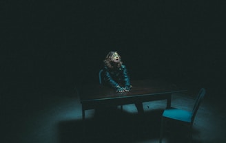 A person with curly hair sits at a wooden table in a dimly lit room, wearing a dark jacket. The atmosphere is tense, with shadows dominating the surroundings. A single chair is placed opposite the person, contributing to an interrogation room-like setting.