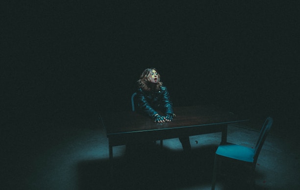 A person with curly hair sits at a wooden table in a dimly lit room, wearing a dark jacket. The atmosphere is tense, with shadows dominating the surroundings. A single chair is placed opposite the person, contributing to an interrogation room-like setting.