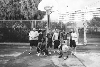 A group of eight men poses on an outdoor basketball court. They appear athletic and are casually dressed, with most of them shirtless. The background includes a basketball hoop, tall palm trees, chain-link fences, and a large building.