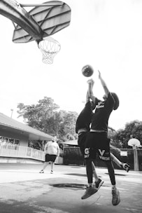 Two young basketball players are jumping to reach for a ball near a basketball hoop. The scene takes place on an outdoor court, with a coach or another individual standing in the background. Trees and a building are visible around the court.