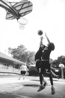 Two young basketball players are jumping to reach for a ball near a basketball hoop. The scene takes place on an outdoor court, with a coach or another individual standing in the background. Trees and a building are visible around the court.