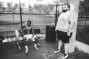 A black and white photo featuring two men in a park setting, with one sitting shirtless on a bench and the other standing beside a trash can. The background includes a metal fence and a soft-focus view of a field with trees.