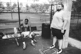A black and white photo featuring two men in a park setting, with one sitting shirtless on a bench and the other standing beside a trash can. The background includes a metal fence and a soft-focus view of a field with trees.