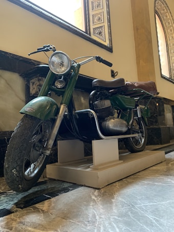 A vintage motorcycle with a green body and a brown leather seat is displayed indoors on a raised platform. The setting has an elegant, museum-like appearance with marble floors and decorative arched windows.