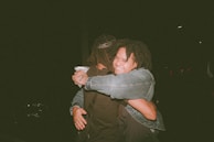 Two people are embracing warmly in a dark setting, where one person is smiling broadly. The scene conveys a sense of warmth and connection.