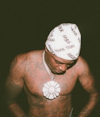 A shirtless man with extensive tattoos on his torso looks downward. He wears a beanie with the word 'NEVER' written multiple times and a thick chain with a large flower-shaped pendant. The photo is taken in low light, emphasizing the textures and details in the tattoos and jewelry.