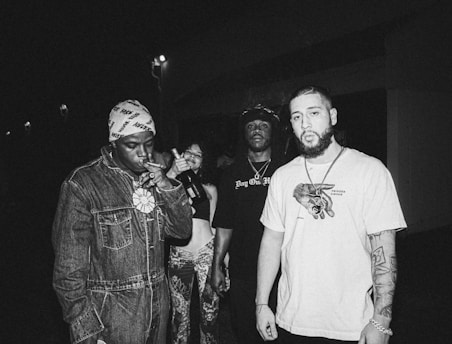 A group of four individuals stand together in a dimly lit environment. One person is smoking a cigar and wearing a beanie and denim jacket, another holds a bottle. A third has long hair and wears a hat and a printed shirt, and the fourth has a beard and tattoos, wearing a T-shirt. The scene appears to be candid and relaxed.