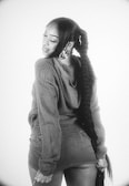 A woman with a long braided hairstyle is posing with her head turned to the side. She is wearing a textured long-sleeve sweater and denim shorts with her hands resting on her thighs. The image is black and white with a plain background.