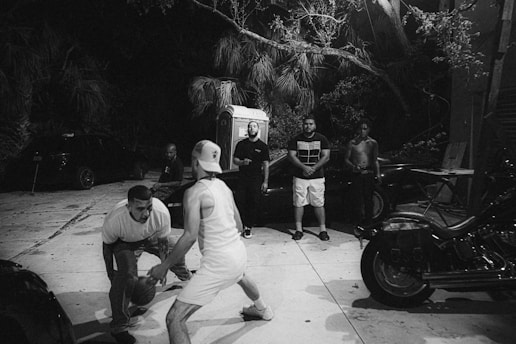 A group of men are gathered outdoors at night, with two of them engaged in a basketball game. There are several spectators watching, some standing and one seated. The setting includes motorcycles, trees, and a portable toilet visible in the background.