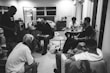 A group of six people are gathered in a room, focusing on music production. One person is working on a computer connected to a large screen displaying music software. Another person is seated beside him with a keyboard, likely for music input. Other individuals are sitting or standing, engaged in conversation, with one person analyzing or working on a small device. The setting appears casual, with a relaxed atmosphere, and the room is furnished with chairs, a table, and a dresser.