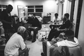 A group of six people are gathered in a room, focusing on music production. One person is working on a computer connected to a large screen displaying music software. Another person is seated beside him with a keyboard, likely for music input. Other individuals are sitting or standing, engaged in conversation, with one person analyzing or working on a small device. The setting appears casual, with a relaxed atmosphere, and the room is furnished with chairs, a table, and a dresser.