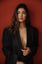 A woman with long, wavy hair wearing an oversized dark blazer against a solid red background. She appears thoughtful, looking slightly to the side with a subtle expression. The blazer is worn loosely, partially revealing her chest.
