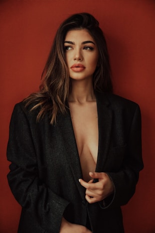 A woman with long, wavy hair wearing an oversized dark blazer against a solid red background. She appears thoughtful, looking slightly to the side with a subtle expression. The blazer is worn loosely, partially revealing her chest.