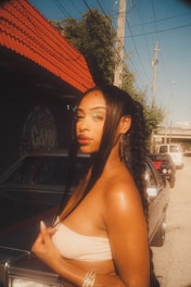 A woman with braided hair stands confidently next to a car on a street. She is wearing earrings and a strapless top, with a backdrop of a building with a red roof and some graffiti art. The scene is sunlit, casting a warm glow over the surroundings.