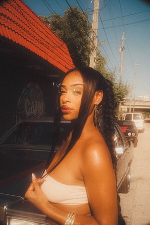 A woman with braided hair stands confidently next to a car on a street. She is wearing earrings and a strapless top, with a backdrop of a building with a red roof and some graffiti art. The scene is sunlit, casting a warm glow over the surroundings.