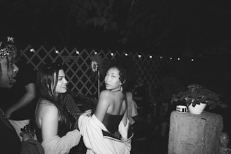 A group of people socializing outdoors at night. A woman in the center is turning towards the camera with a slight smile, while others around her are engaged in conversation. String lights are hung in the background over a lattice fence, creating a cozy ambiance. A plant in a pot is placed on a concrete pillar in the foreground.