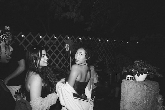 A group of people socializing outdoors at night. A woman in the center is turning towards the camera with a slight smile, while others around her are engaged in conversation. String lights are hung in the background over a lattice fence, creating a cozy ambiance. A plant in a pot is placed on a concrete pillar in the foreground.