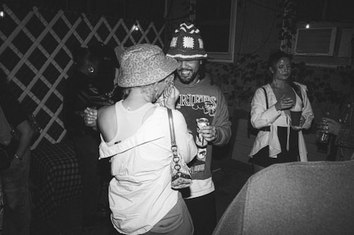 A black-and-white photograph depicting a group of people socializing at a party. One person is wearing a patterned bucket hat and tank top, dancing closely with another wearing a beanie and graphic sweater, holding a drink can. Other attendees are visible in the background, holding cups and appearing engaged in conversation. The setting suggests an outdoor night gathering, possibly in a backyard, with ambient lighting and ivy-covered walls.