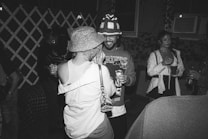 A black-and-white photograph depicting a group of people socializing at a party. One person is wearing a patterned bucket hat and tank top, dancing closely with another wearing a beanie and graphic sweater, holding a drink can. Other attendees are visible in the background, holding cups and appearing engaged in conversation. The setting suggests an outdoor night gathering, possibly in a backyard, with ambient lighting and ivy-covered walls.