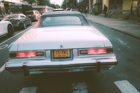 A classic white Buick car is parked on an urban street, with a New York license plate. The scene includes a few other parked cars, a sidewalk, and buildings with pedestrians in the background. The lighting gives a nostalgic and serene mood.