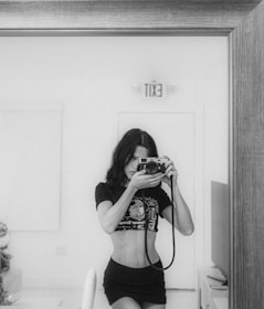 A black and white photograph features a person with long hair holding a camera in front of a mirror, capturing their own image. The individual is wearing a crop top and a skirt. The room appears simple and minimalistic, with an 'Exit' sign reflected in the mirror above the person’s head.