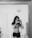 A black and white photograph features a person with long hair holding a camera in front of a mirror, capturing their own image. The individual is wearing a crop top and a skirt. The room appears simple and minimalistic, with an 'Exit' sign reflected in the mirror above the person’s head.