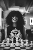 A person with curly hair is smiling while sitting at a chessboard. They are wearing a sleeveless shirt with 'Brooklyn Original Vintage' printed on it. The scene is in black and white, giving it a timeless or retro feel, and is set indoors with a blurred backdrop.