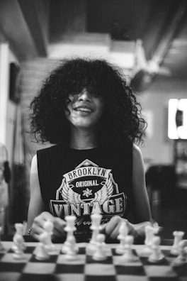 A person with curly hair is smiling while sitting at a chessboard. They are wearing a sleeveless shirt with 'Brooklyn Original Vintage' printed on it. The scene is in black and white, giving it a timeless or retro feel, and is set indoors with a blurred backdrop.