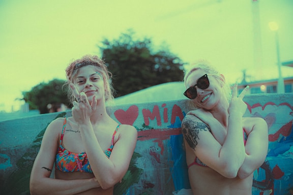 Two individuals are standing in front of a graffiti-covered wall. They are both wearing colorful bikinis and are posing with crossed arms, showing off their tattoos and smiling. One of them is wearing sunglasses. The wall has a heart symbol and the word 'MIA' painted in red.