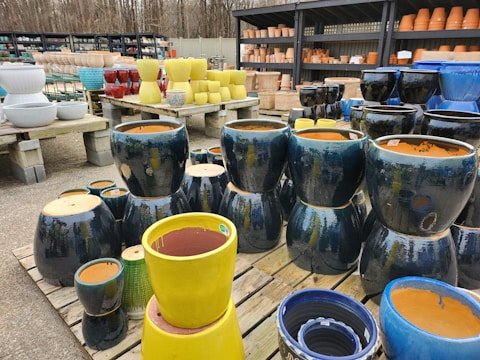 Various ceramic pots of different sizes and colors are displayed on wooden pallets. The pots have glossy finishes, with colors including blue, yellow, red, and white. The background shows shelves filled with more pots against a fence and bare trees.