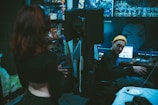 A recording studio with a dim, moody atmosphere. One person stands in the foreground with their back to the camera, while another person wearing a yellow beanie is seated at a desk with audio equipment. The walls are decorated with acoustic foam panels and anime posters. A large computer screen displays an anime character's image. Some clutter, such as cables and a plate with food, is visible on the desk.