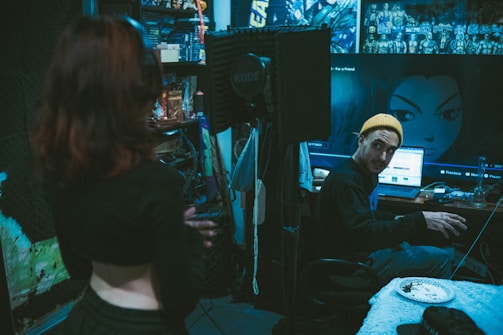 A recording studio with a dim, moody atmosphere. One person stands in the foreground with their back to the camera, while another person wearing a yellow beanie is seated at a desk with audio equipment. The walls are decorated with acoustic foam panels and anime posters. A large computer screen displays an anime character's image. Some clutter, such as cables and a plate with food, is visible on the desk.