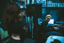 A recording studio with a dim, moody atmosphere. One person stands in the foreground with their back to the camera, while another person wearing a yellow beanie is seated at a desk with audio equipment. The walls are decorated with acoustic foam panels and anime posters. A large computer screen displays an anime character's image. Some clutter, such as cables and a plate with food, is visible on the desk.