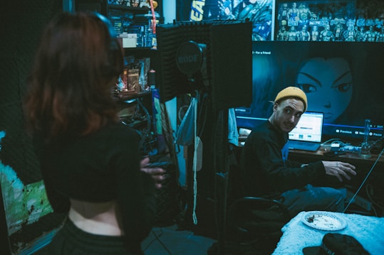 A recording studio with a dim, moody atmosphere. One person stands in the foreground with their back to the camera, while another person wearing a yellow beanie is seated at a desk with audio equipment. The walls are decorated with acoustic foam panels and anime posters. A large computer screen displays an anime character's image. Some clutter, such as cables and a plate with food, is visible on the desk.