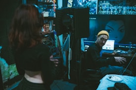 A recording studio with a dim, moody atmosphere. One person stands in the foreground with their back to the camera, while another person wearing a yellow beanie is seated at a desk with audio equipment. The walls are decorated with acoustic foam panels and anime posters. A large computer screen displays an anime character's image. Some clutter, such as cables and a plate with food, is visible on the desk.