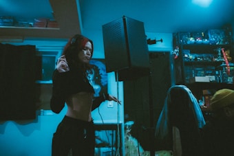 A young woman with partially dyed hair is in a recording studio, standing near a microphone with a pop filter. The room is dimly lit with blue lighting, and various items, including shelves filled with collectibles and books, are visible in the background. Another person is sitting at a desk, wearing a yellow beanie.