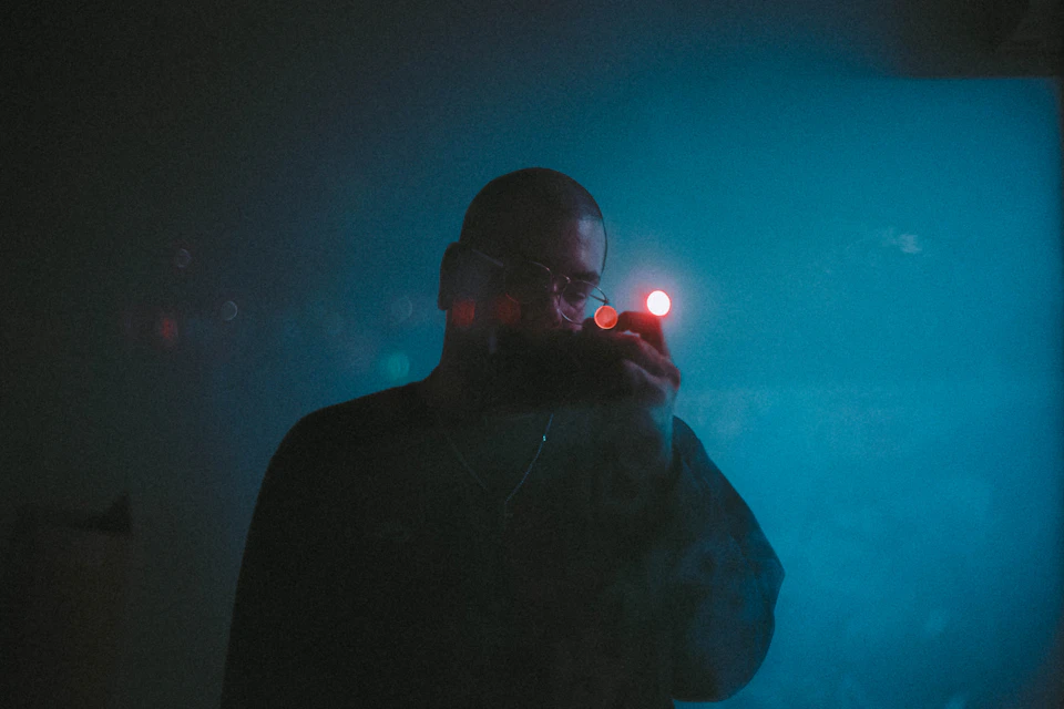 The image shows a man standing against a blue wall, illuminated by red lights that cast an ethereal glow on his face and upper body. The man is wearing glasses and has a beard, adding to his mysterious appearance. He holds a small object in his hand, which appears to be a camera or a phone.
