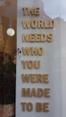 Large gold letters are arranged vertically on a white background, spelling out an inspirational message. A simple and modern style is evident, positioned in or near a window display, with a reflection of buildings. Red berries and branches are in a woven basket at the base.