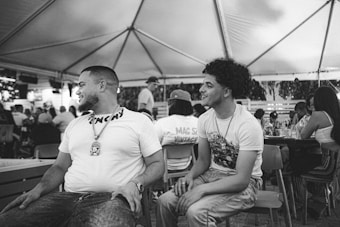 Two men are sitting side by side under a large tent, engaging in conversation. They are surrounded by other people seated at tables, indicating a social gathering or event. The setting has an open and relaxed atmosphere.
