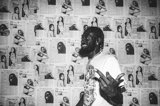 A man smiles broadly, standing against a background of newspaper clippings featuring various images and articles. The clippings are predominantly black and white, creating a collage effect on the wall behind him. The man is wearing a patterned shirt and has an expressive hand gesture.