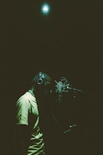 A person wearing headphones is standing in front of a microphone in a dimly lit recording studio. The lighting creates a soft, atmospheric mood with a focus on the person and the microphone. A bright light is visible at the top of the image, illuminating the scene with a greenish hue.