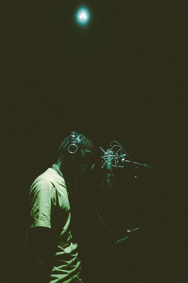 A person wearing headphones is standing in front of a microphone in a dimly lit recording studio. The lighting creates a soft, atmospheric mood with a focus on the person and the microphone. A bright light is visible at the top of the image, illuminating the scene with a greenish hue.