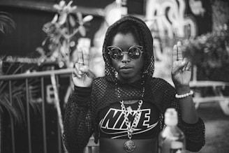 A person wearing round sunglasses and a netted top poses with both hands raised in a peace sign. The individual accessorizes with a nose ring, a large medallion necklace, and several bracelets. The background features graffiti art and blurred foliage.