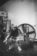 A person with long curly hair and wearing casual clothing, including a headband, crouches in front of a large industrial fan. The setting appears to be an indoor space with a rugged, artistic vibe, featuring various items like frames and plants in the background.