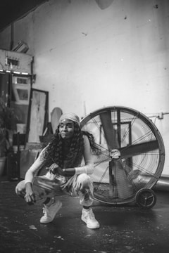A person with long curly hair and wearing casual clothing, including a headband, crouches in front of a large industrial fan. The setting appears to be an indoor space with a rugged, artistic vibe, featuring various items like frames and plants in the background.
