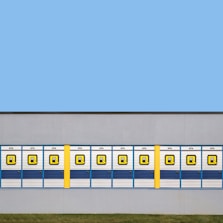 A row of symmetrical blue and white garage doors with a bold yellow column interrupting the pattern, set against a flat gray wall. The top half of the image features a clear blue sky, while a strip of green grass lies at the bottom.
