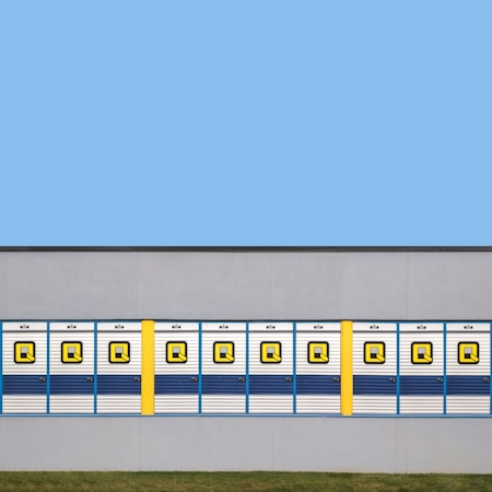 A row of symmetrical blue and white garage doors with a bold yellow column interrupting the pattern, set against a flat gray wall. The top half of the image features a clear blue sky, while a strip of green grass lies at the bottom.
